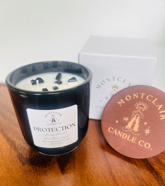 A black candle labeled 'PROTECTION' with crystals on top, placed on a wooden surface with cover and box