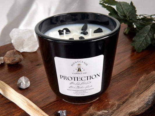 A black candle labeled 'PROTECTION' with crystals on top, placed on a wooden surface with a stick of palo santo, crystals, and a white candle in the background.