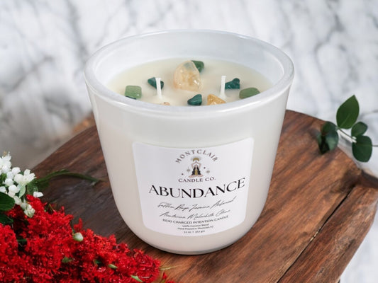 A white candle labeled 'ABUNDANCE' with crystals on top, placed on a wooden surface, surrounded by red and white flowers.