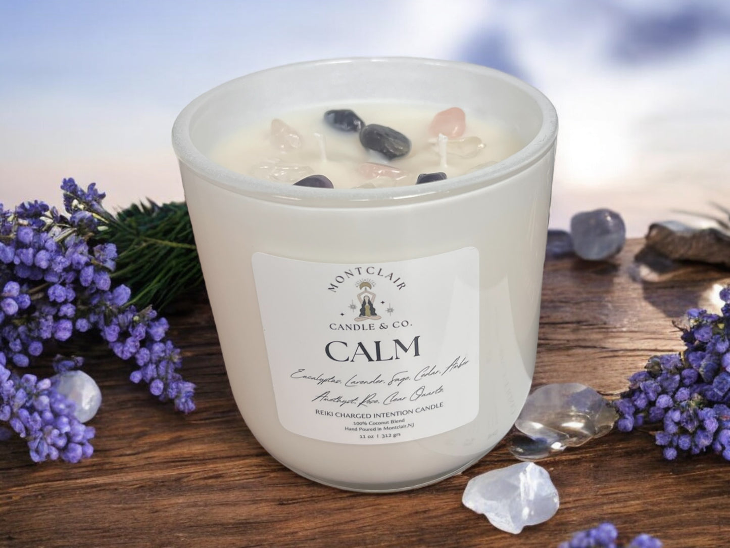 A white candle with crystals on top, labeled 'CALM', surrounded by purple flowers and charging stones on a wooden surface with a blurred background.