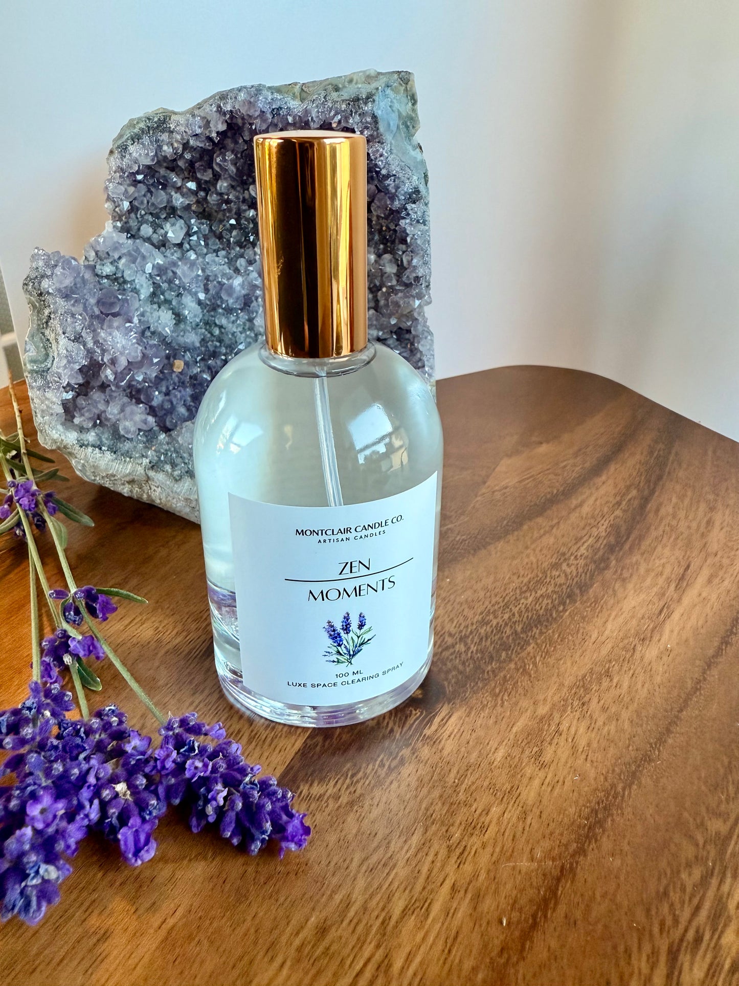 Bottle of 'Zen Moments' perfume with a gold cap on a wooden surface with lavender flowers and a crystal.