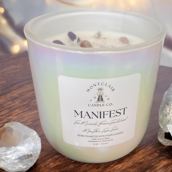 Zoomed in white candle with the word 'MANIFEST' written on the label, set on a wooden surface with crystals around it.