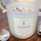 Zoomed in white candle with the word 'MANIFEST' written on the label, set on a wooden surface with crystals around it.