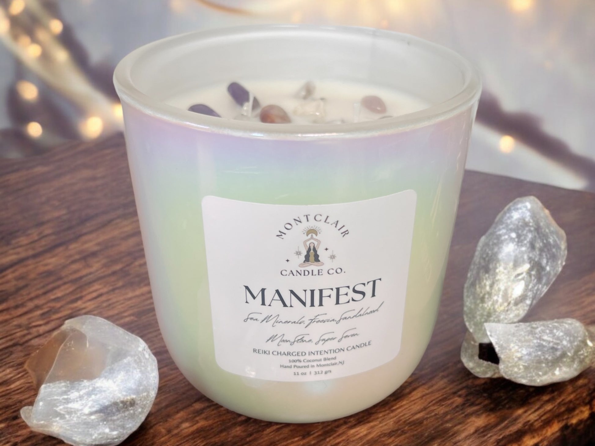 A white candle with the word 'MANIFEST' written on the label, set on a wooden surface with crystals around it.