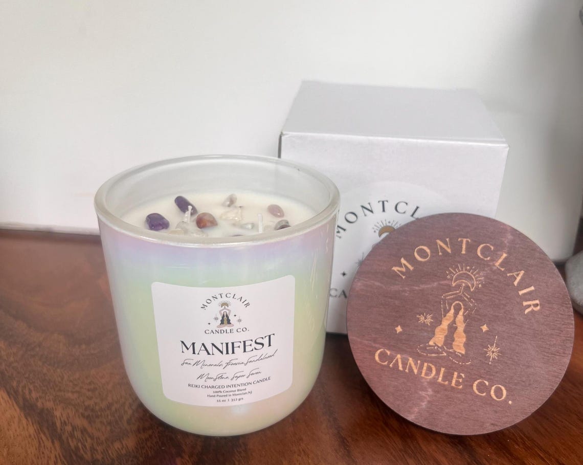 A white candle with the word 'MANIFEST' written on the label, set on a wooden surface with cover and box