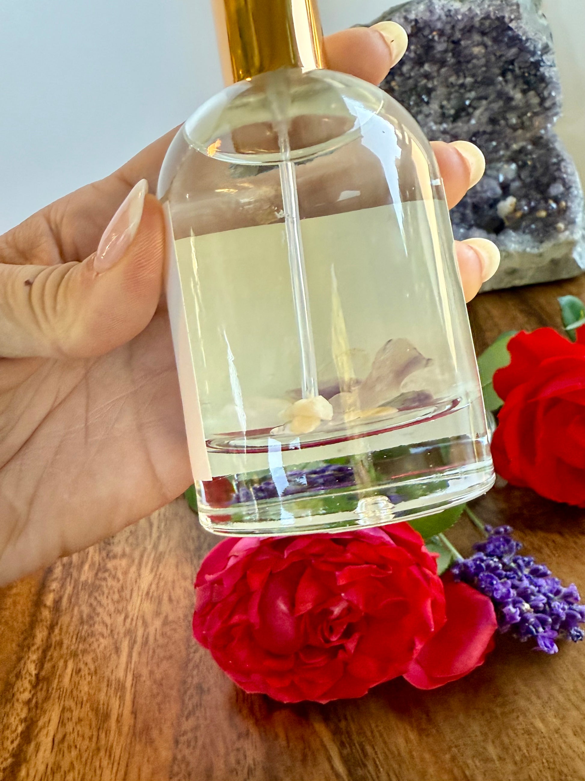 Clear bottle with gold cap held by a hand, surrounded by flowers on a wooden surface.
